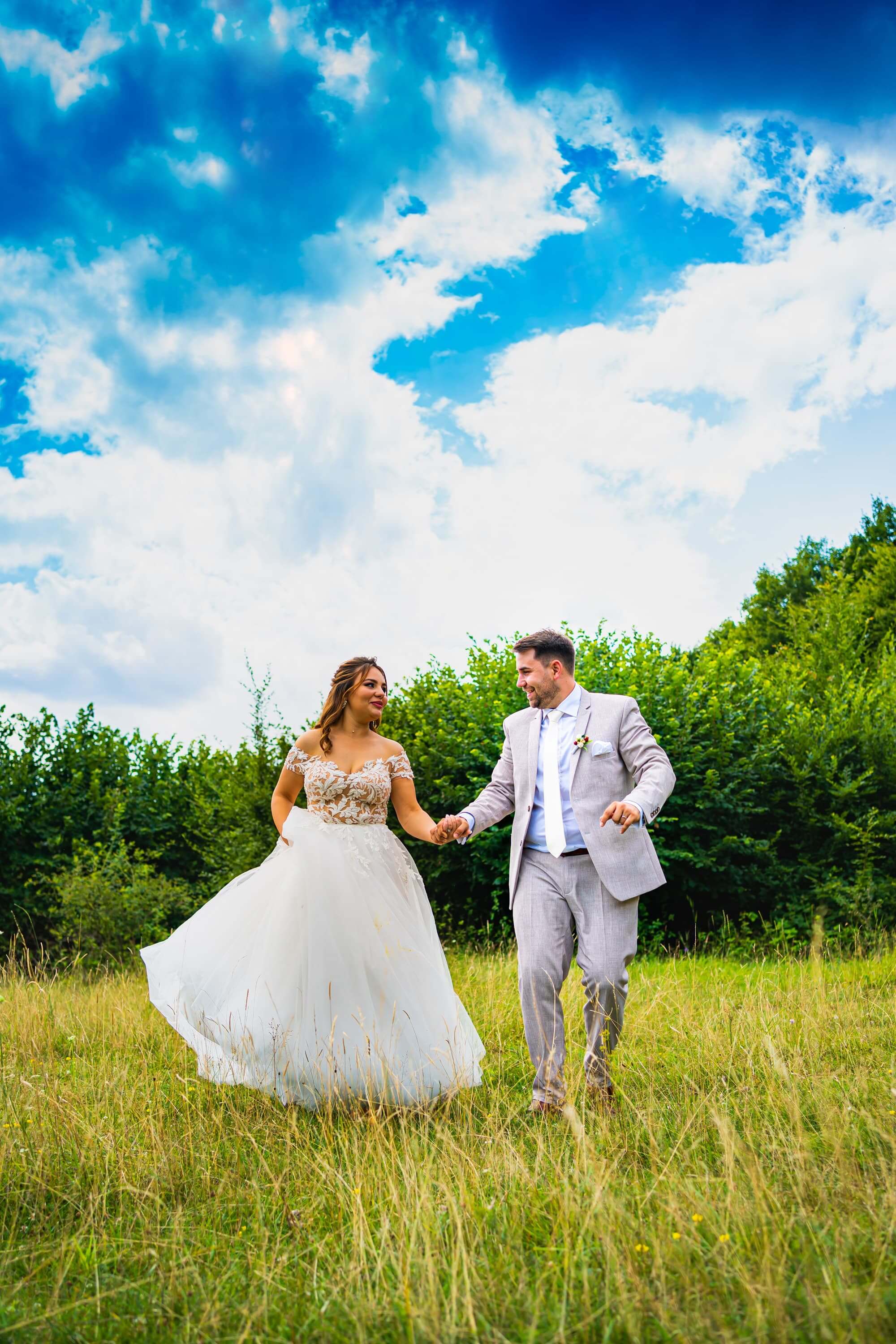 Fotografie de nuntă realizată de Flaviu Studio cu Anca și Paul plimbându-se mână în mână într-o pajiște verde sub un cer senin. Mireasa poartă o rochie albă cu dantelă, iar mirele este îmbrăcat într-un costum deschis la culoare. Fundalul natural creează o atmosferă romantică și liberă.