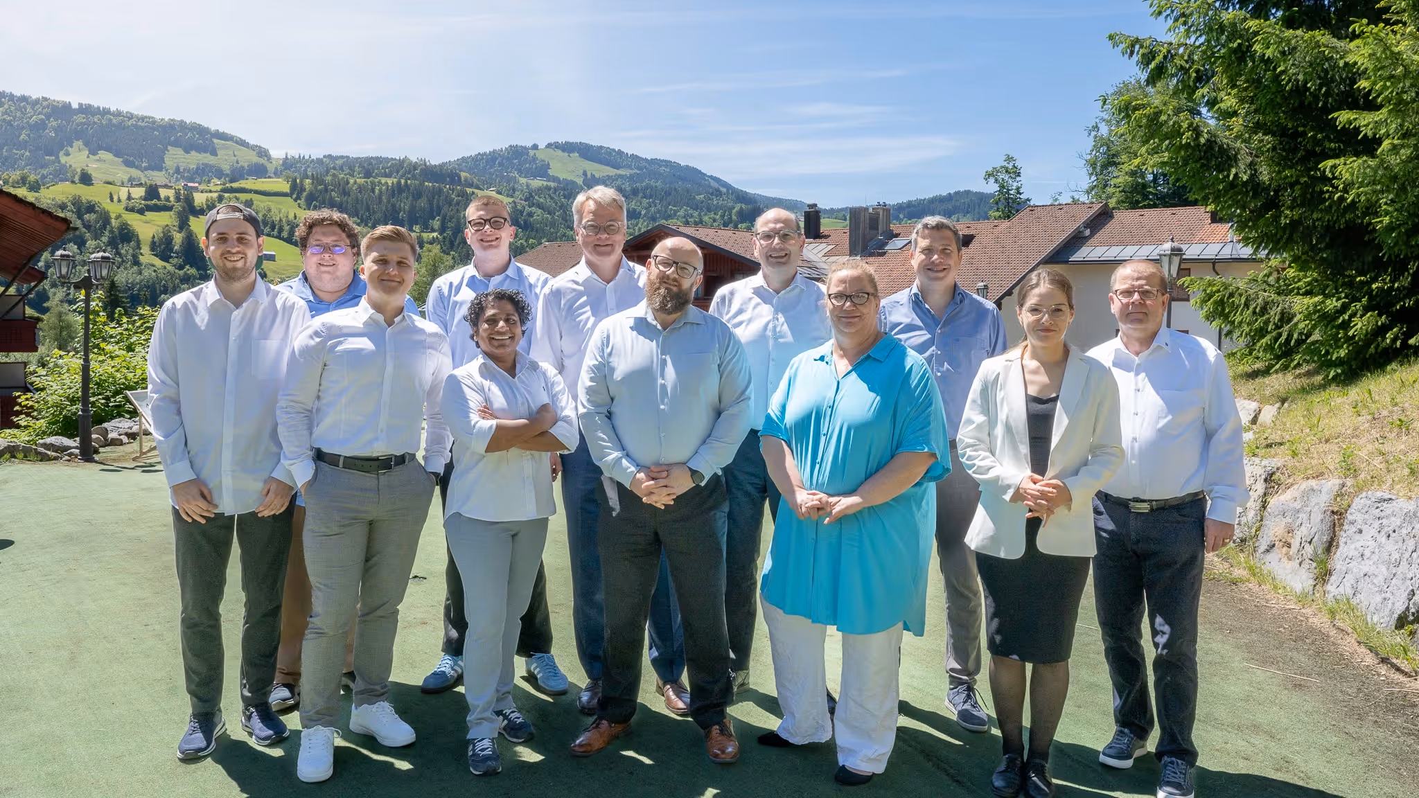 Gruppenfoto von zwölf Personen, die bei sonnigem Wetter draußen mit Bergen und Häusern im Hintergrund stehen.