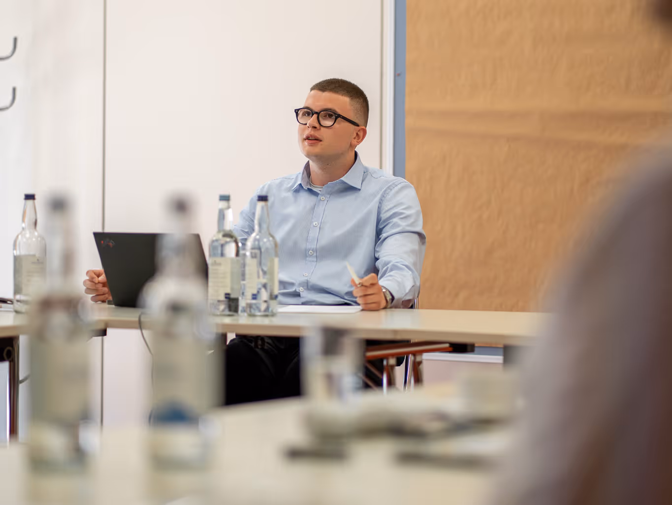 Junger Mann mit Brille spricht in einem Konferenzraum mit Wasserflaschen auf dem Tisch.