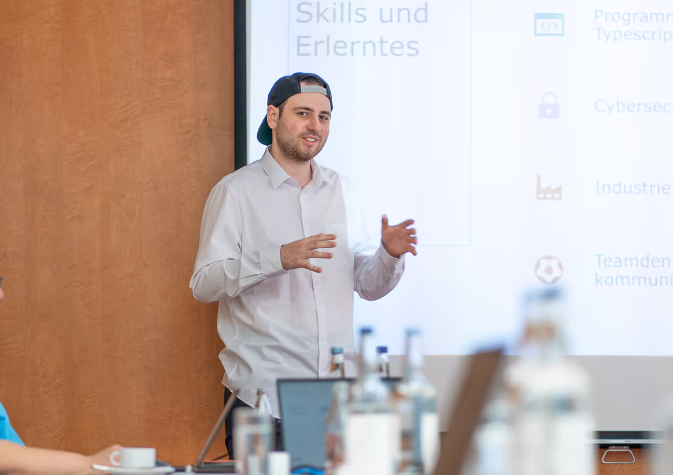 Mann in weißem Hemd und rückwärts getragenem Cap gibt Präsentation vor Publikum mit Laptop und Wasserflaschen.