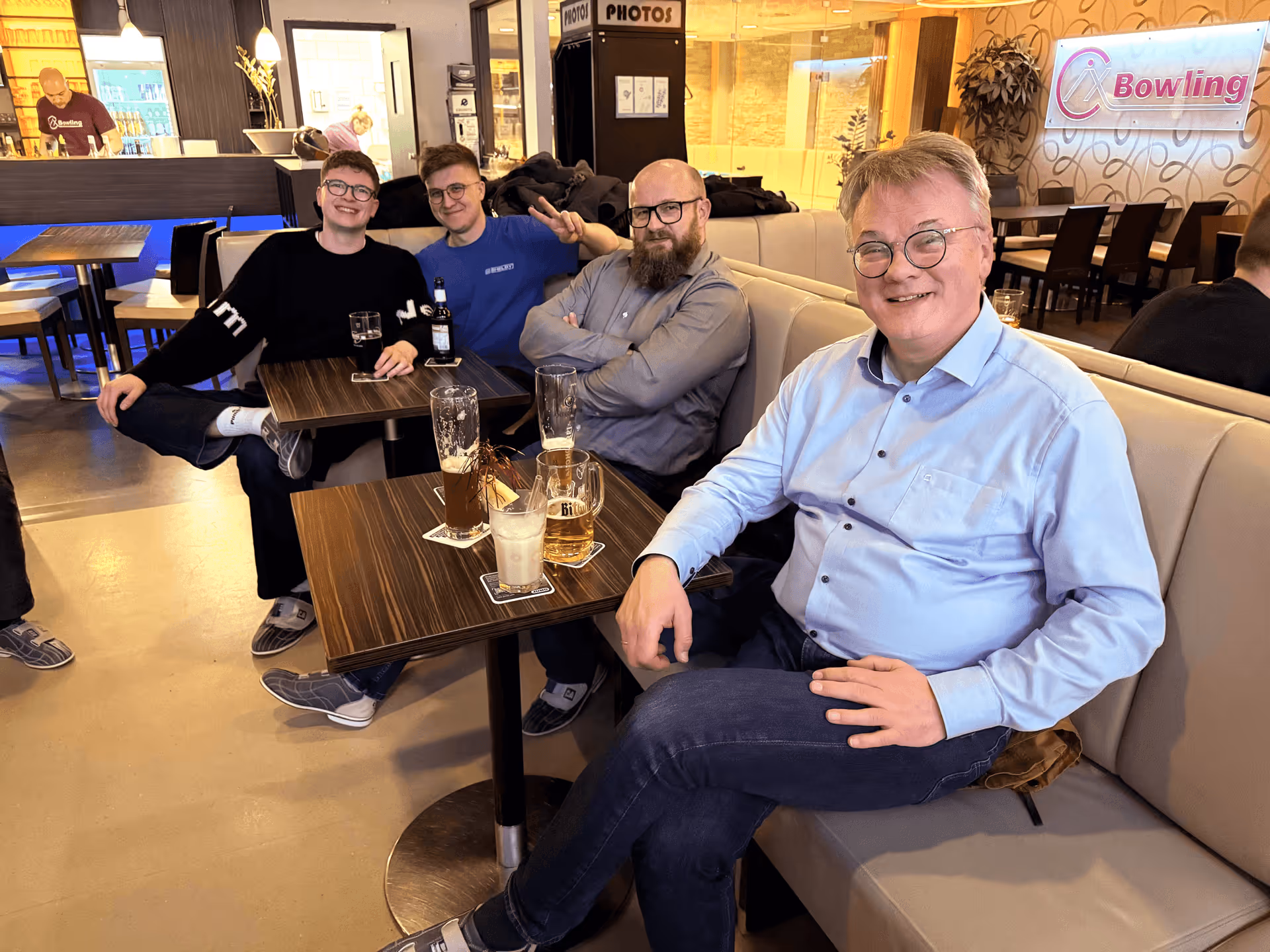 Vier Männer sitzen in einer Bowlingbahn-Lounge an Tischen mit Getränken, lächelnd und entspannt.