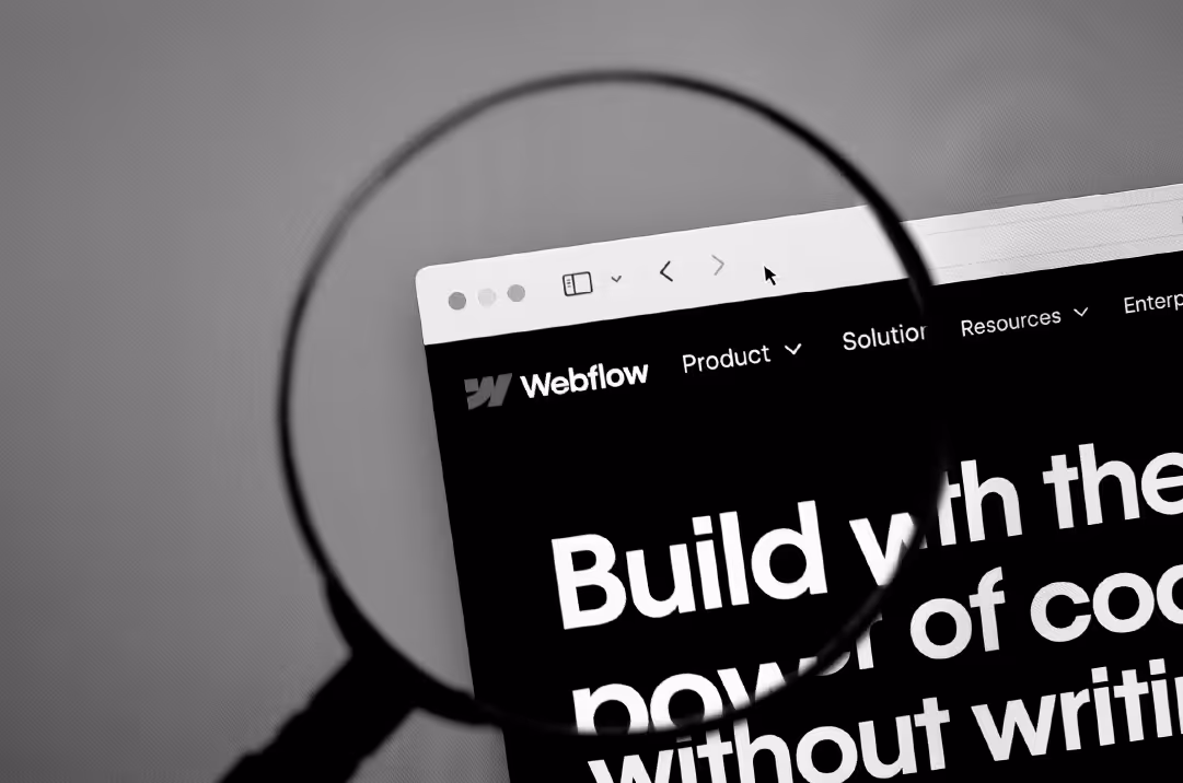 A magnifying glass is held over a computer screen displaying the Webflow homepage.