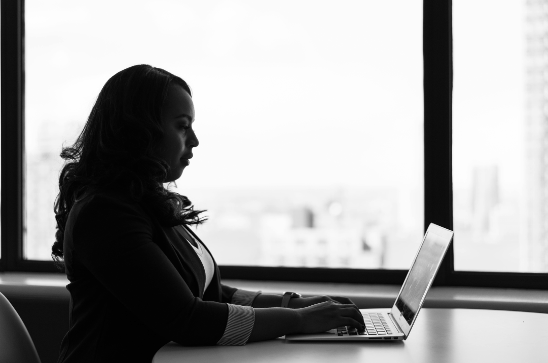 A woman works on her laptop in an office with a large window.