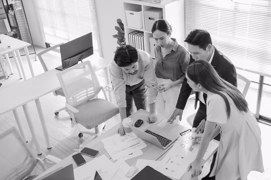 A team of men and women gather around a laptop on a table in an office setting.