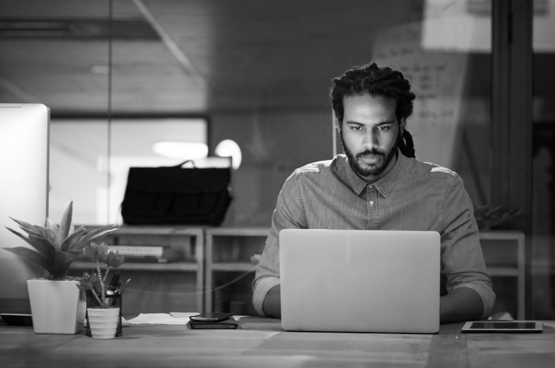 A man with locs and a sweater sits behind a laptop working, his eyes focused on the screen.