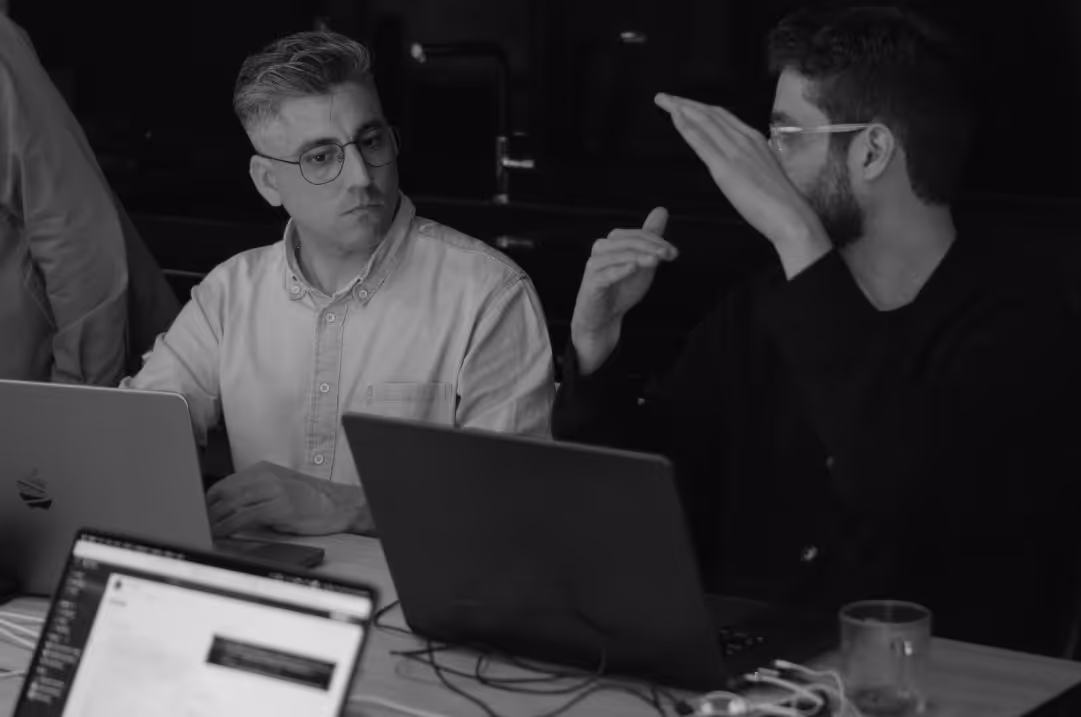 Two men face each other during a conversation held as they sit next to each other in front of their laptops.