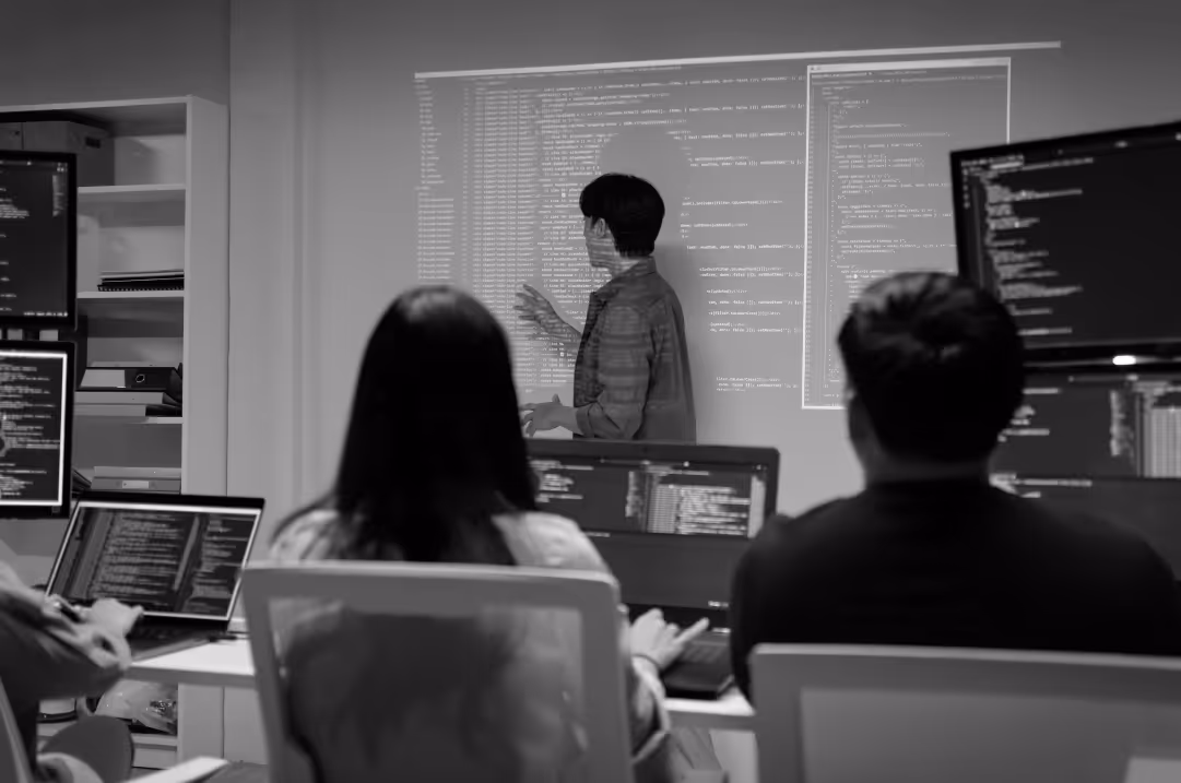 An instructor stands at the front of a room reviewing code while students sit in front of their own computers, which also have code on the screens.