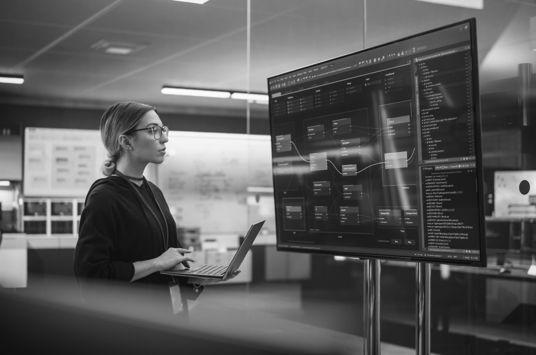 A woman stands holding her laptop while gazing at the screen of a large monitor.