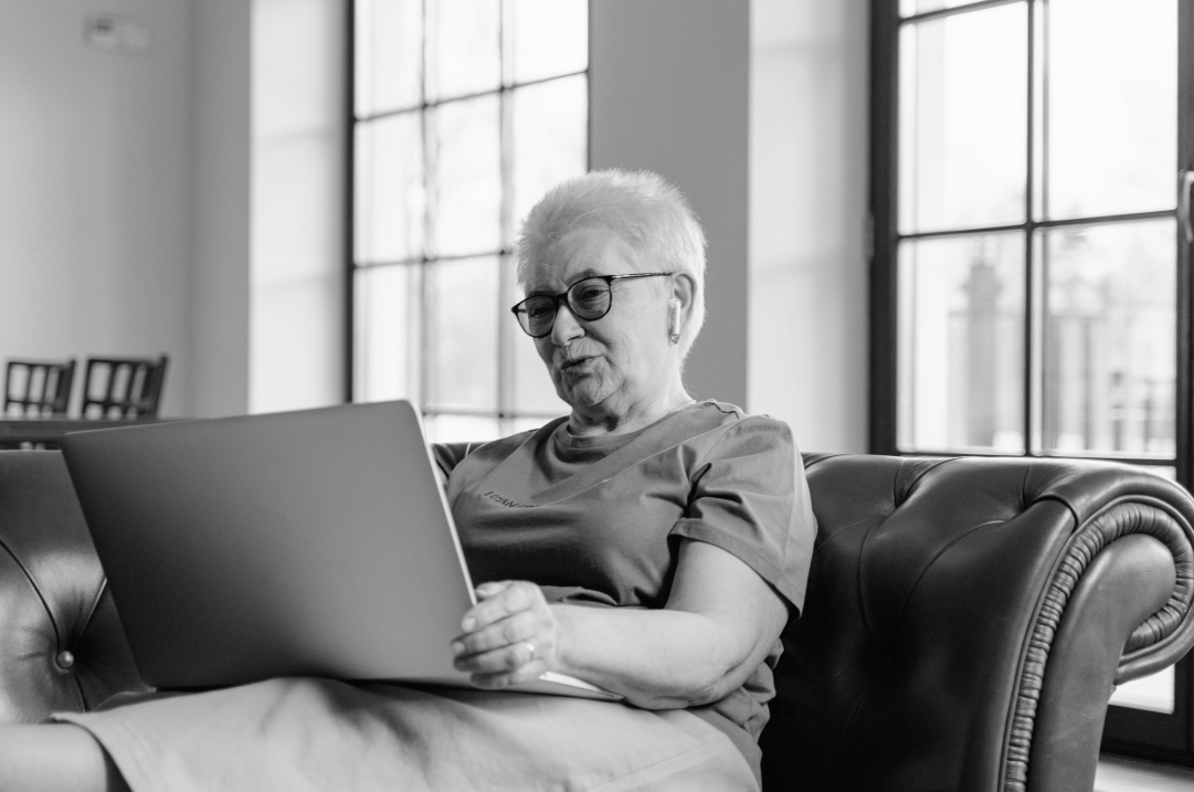 An older woman sits on their couch, smiling as they look at the laptop on their lap.