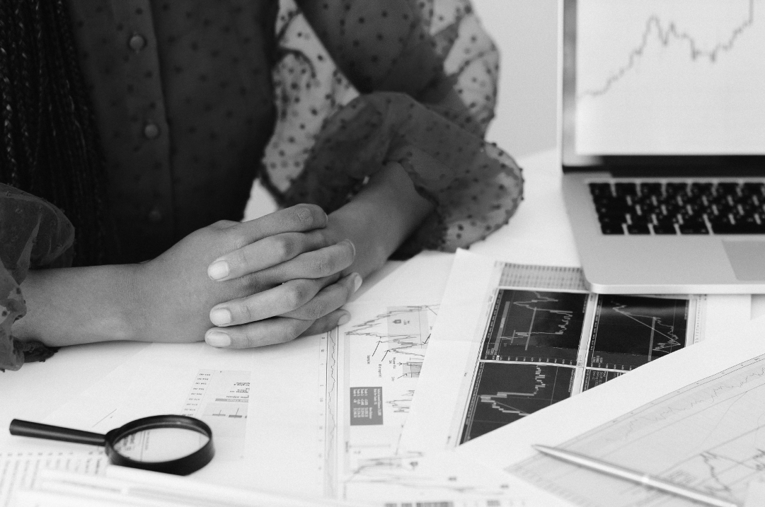 A woman sits with her hands folded on her desk, which contains charts and her laptop.