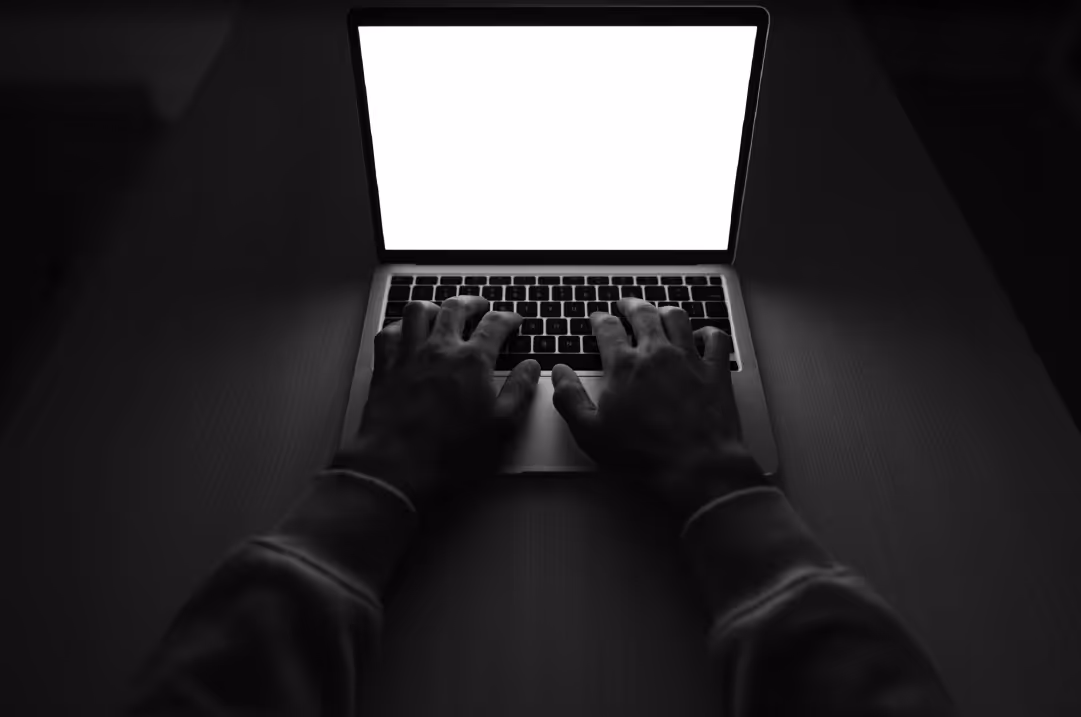 A person in a dark room rests their hands on the keyboard of a glowing laptop.