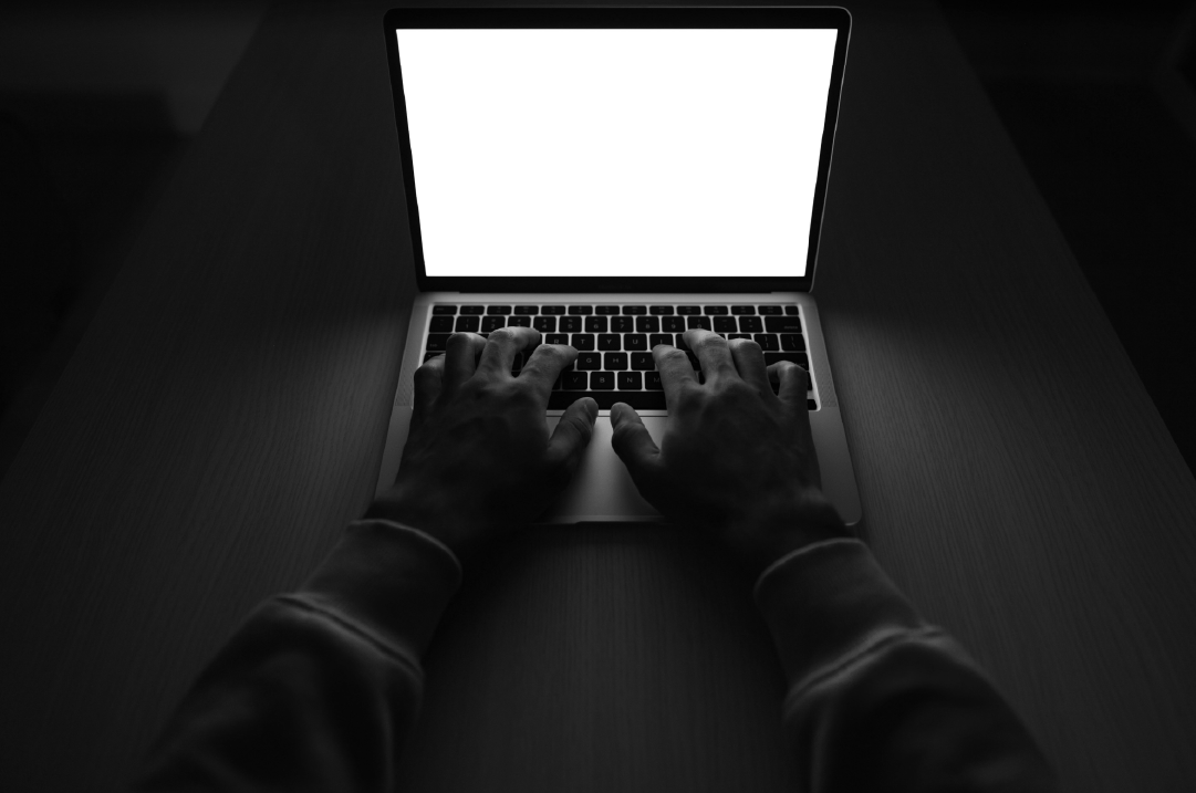 A person in a dark room rests their hands on the keyboard of a glowing laptop.