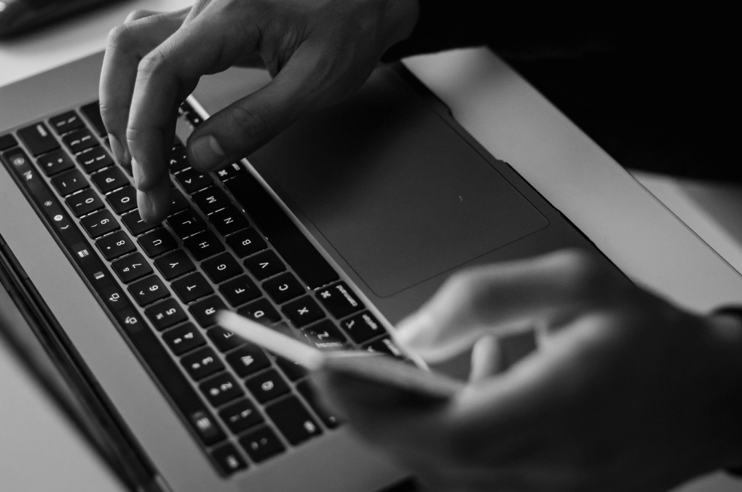 A person types on their laptop with one hand while holding a mobile phone in their other hand.