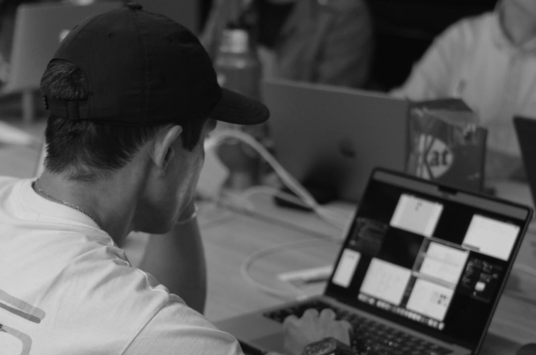 A person wearing a baseball cap is looking at a laptop while seated in a workspace with other people.