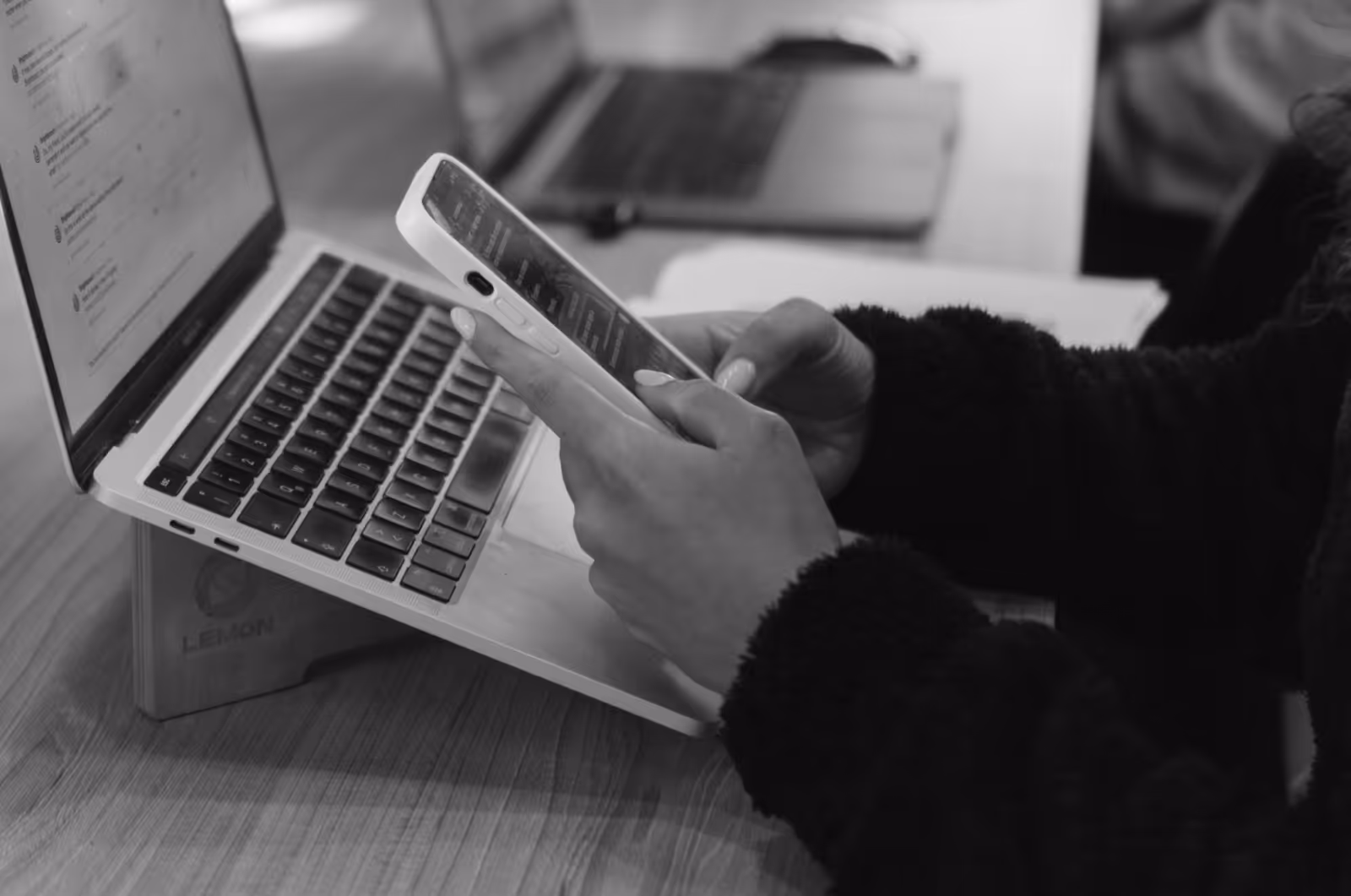 A woman is typing on her mobile phone. An open laptop rests in front of her on her desk.