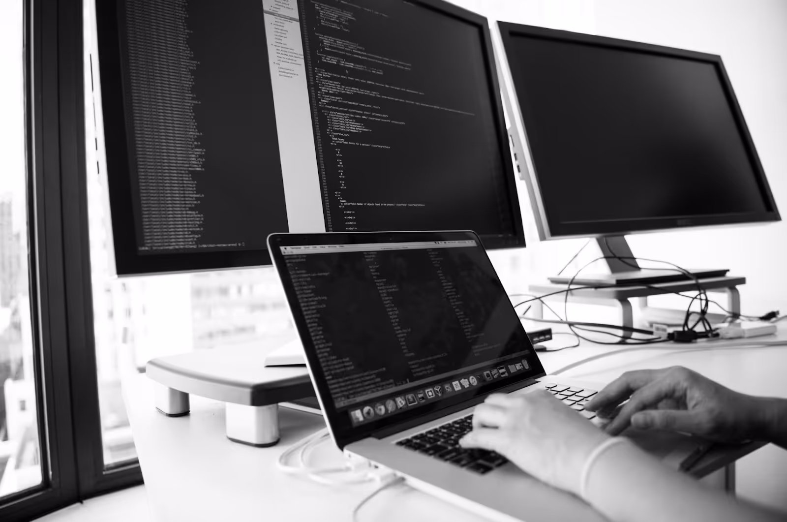 Hands type on a laptop that rests on a desk with three other monitors.
