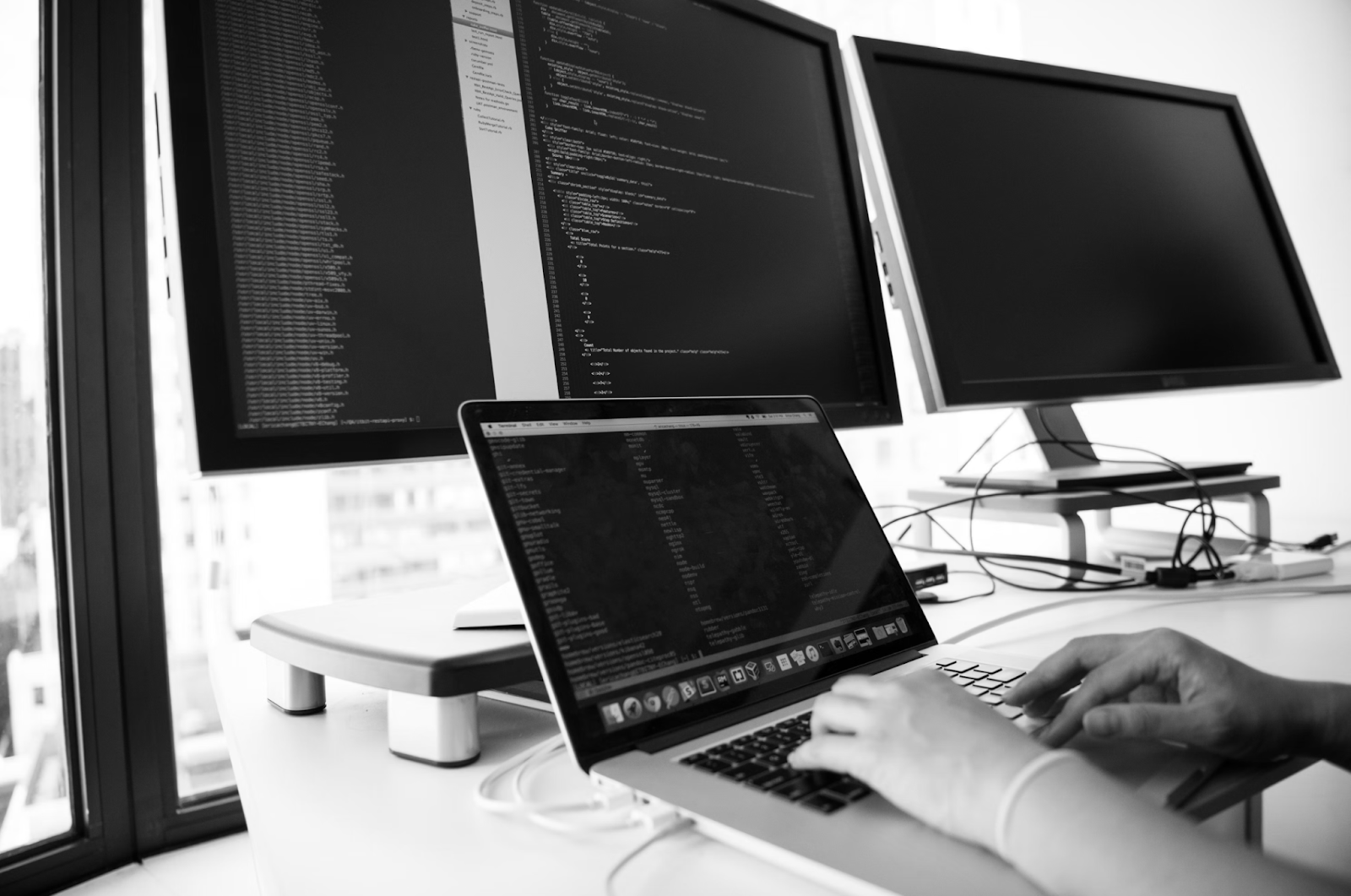 Hands type on a laptop that rests on a desk with three other monitors.