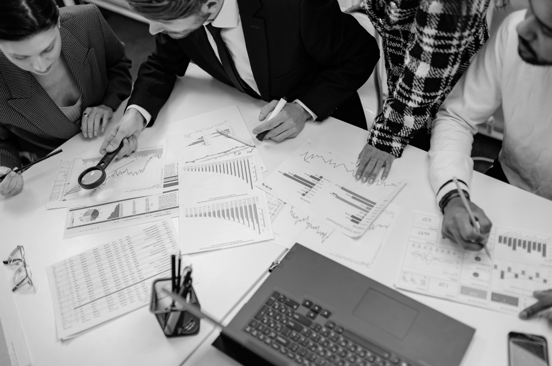 Several people are gathered around a table in an office, some sitting and others standing, looking at documents that contain different graphs and charts.