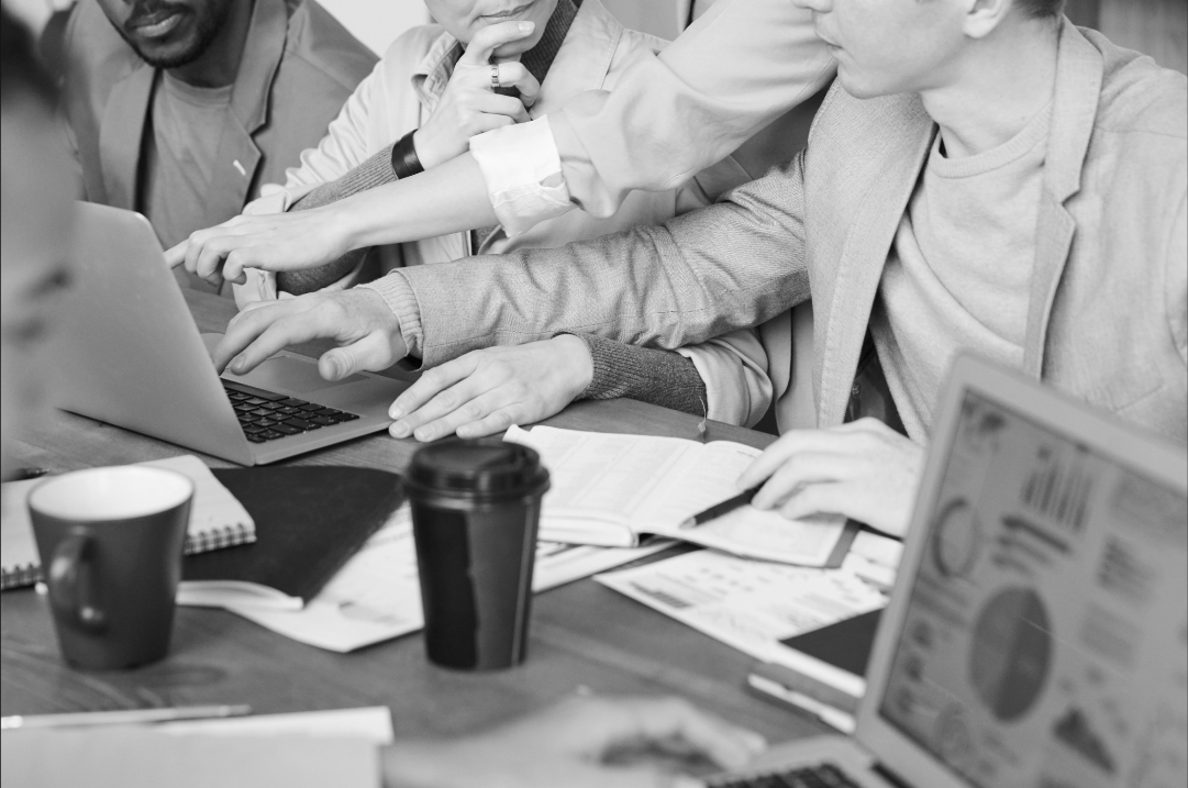 Several people are gathered around a table, two of them gesturing at a laptop, while others in attendance pay attention.