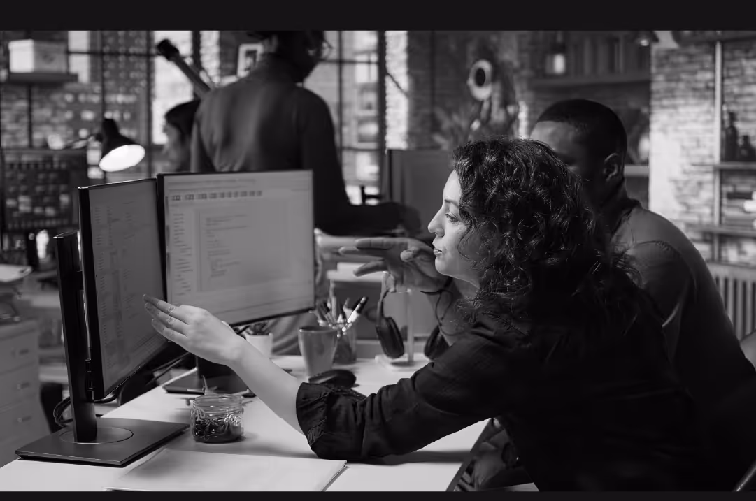 A woman and a man work together in an office, looking at two monitors.