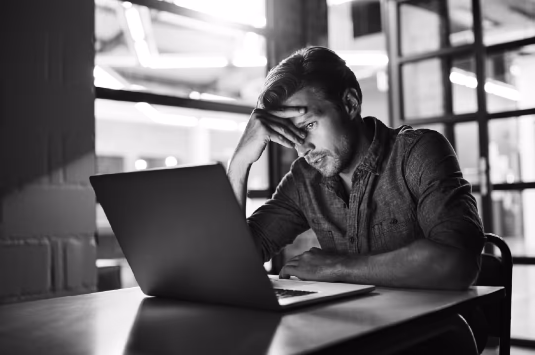 A man rests his head on his hand while peering at a laptop with a perplexed expression.