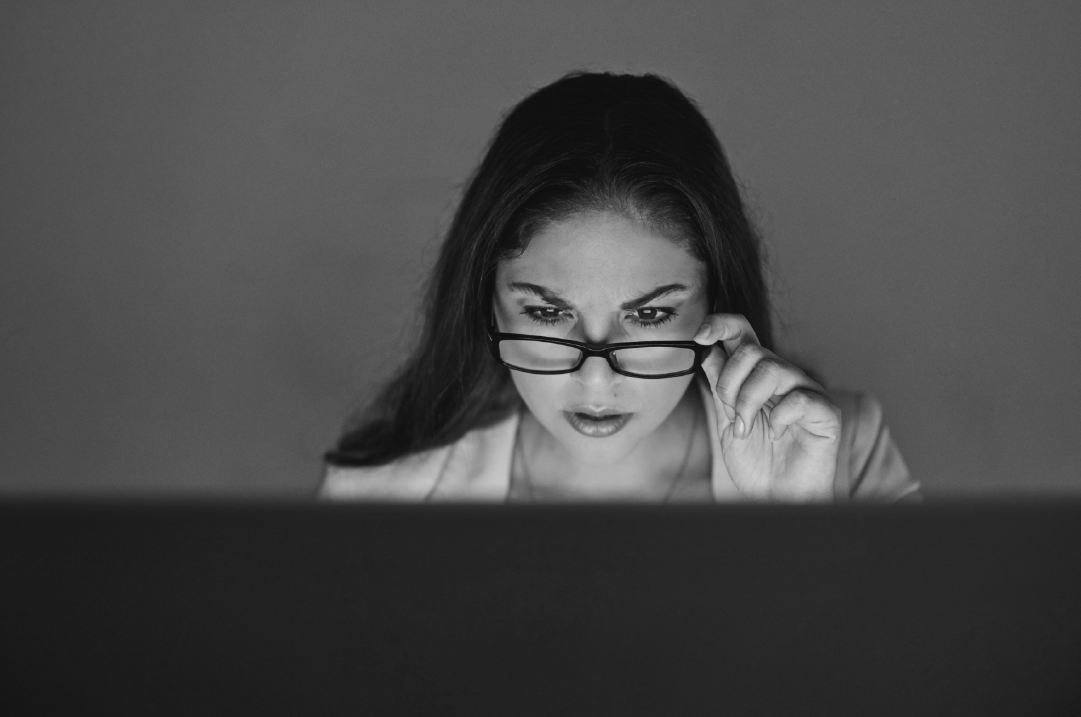 A woman peers over her glasses, staring at something with a shocked expression.