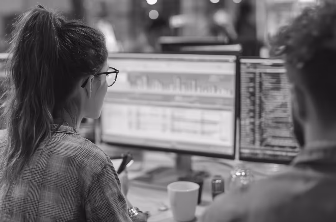 Two software developers working at dual monitors in an office, analyzing data dashboards and writing code.