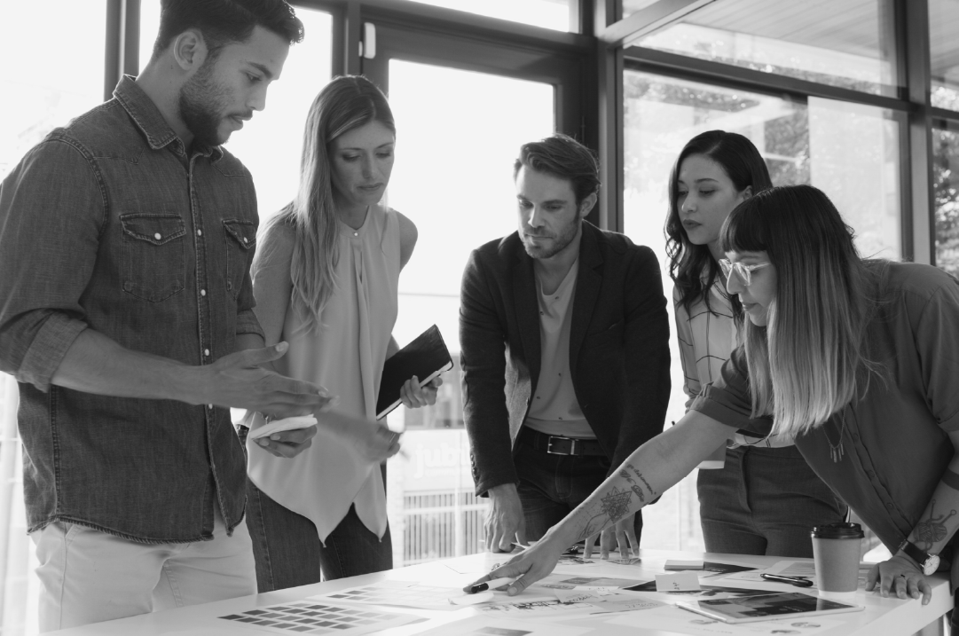 Five people stand around a table in an office setting reviewing documents.
