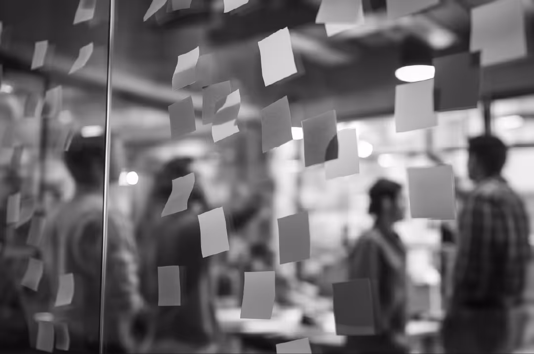 A wall of sticky notes in the foreground on a glass surface in an office setting. People in the background are standing around office equipment.