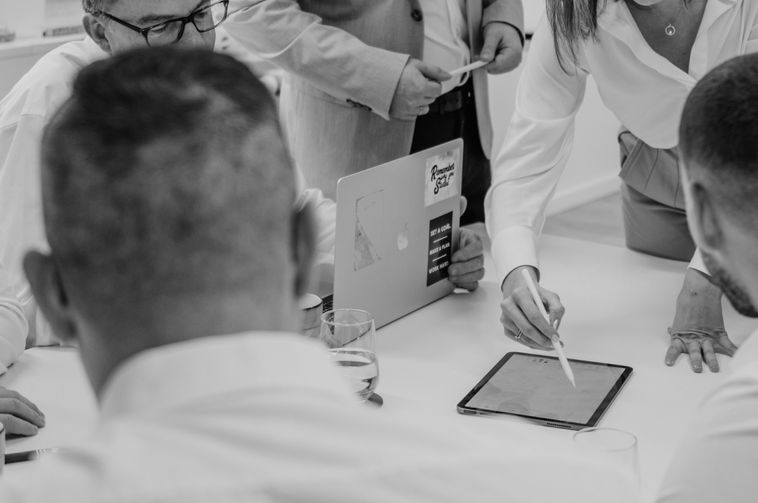 People stand around a table in an office looking at a laptop and a tablet.