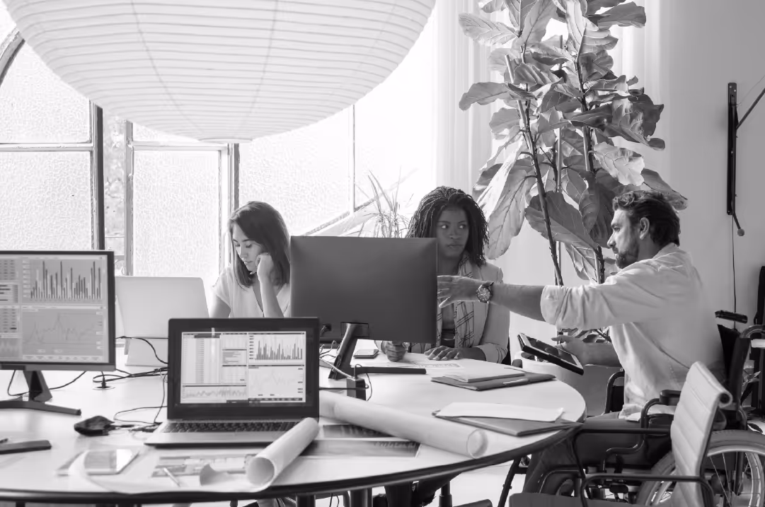 Three individuals sit around a table containing multiple laptops and monitors in a spacious office setting. 