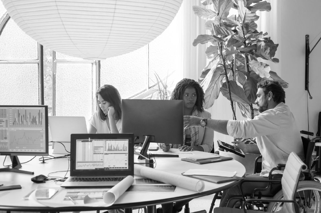 Three individuals sit around a table containing multiple laptops and monitors in a spacious office setting. 