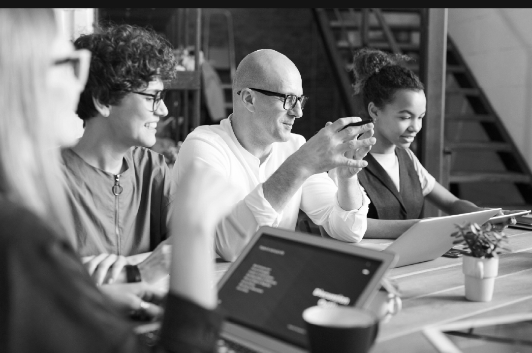 Four individuals smile while seated next to each other and working on their laptops.
