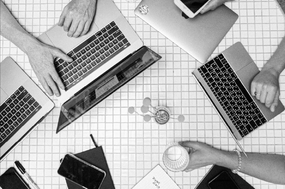 An overhead shot of multiple laptops and cellphones on a table surrounded by hands.