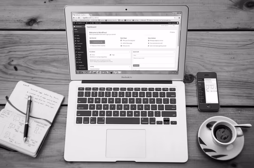 A laptop sits on a table displaying the backend of Wordpress. A notebook, a cell phone, and a cup of coffee are also on the table.