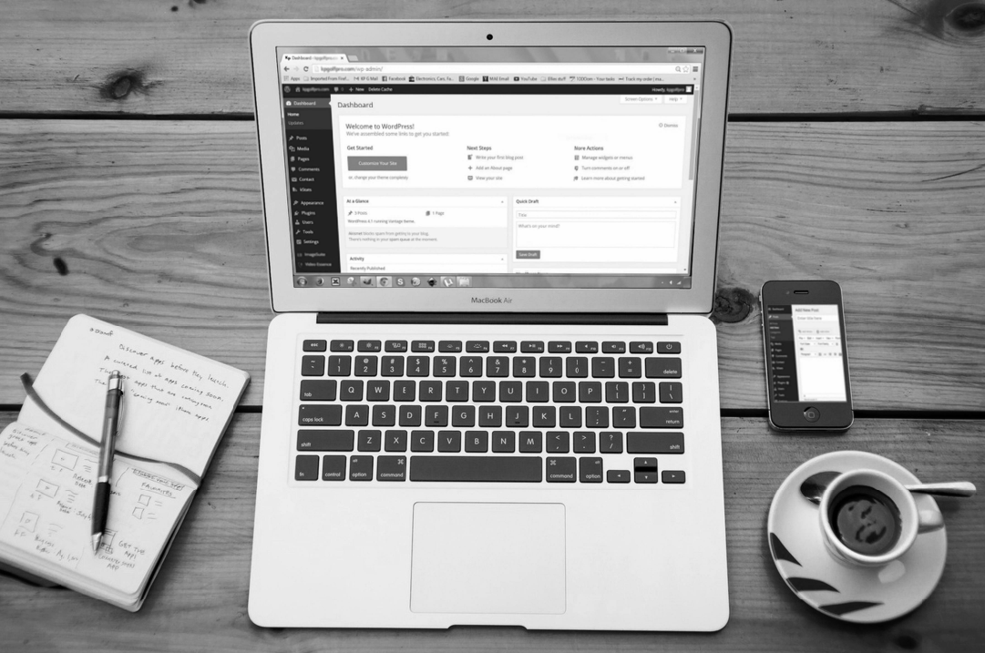A laptop sits on a table displaying the backend of Wordpress. A notebook, a cell phone, and a cup of coffee are also on the table.