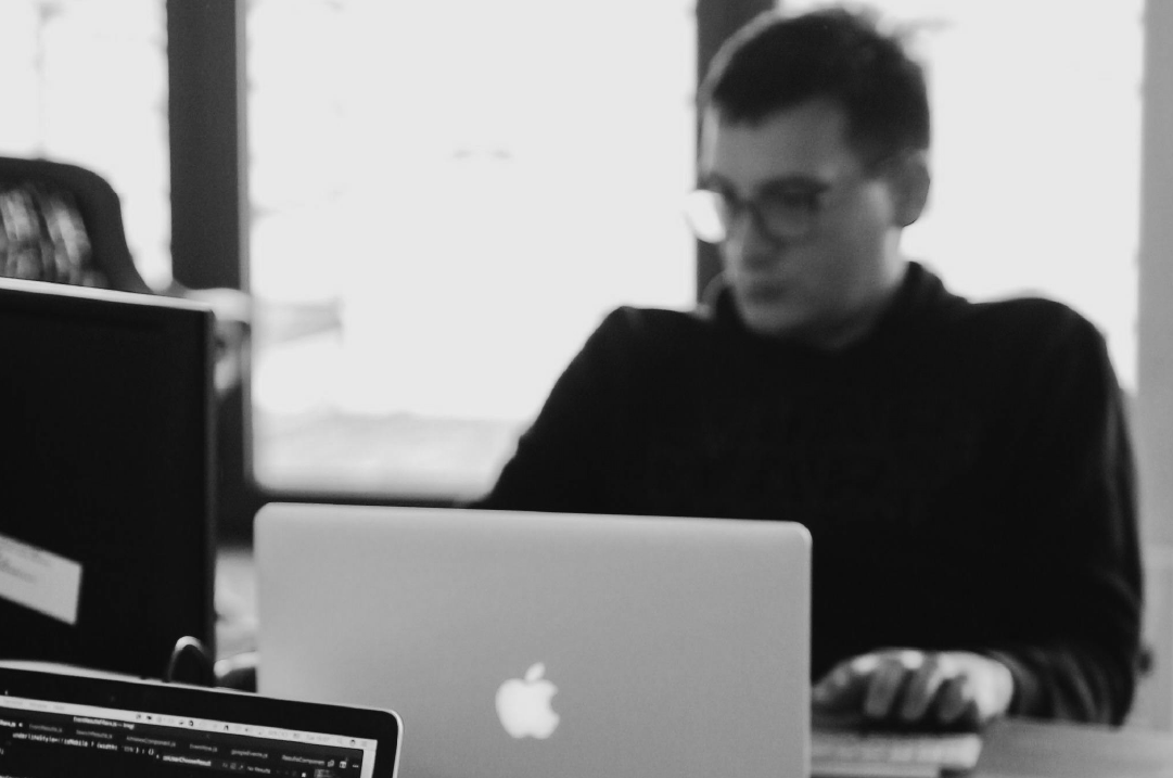 A man in a black turtleneck is in a workspace with a laptop and a monitor on the desk where he is seated.