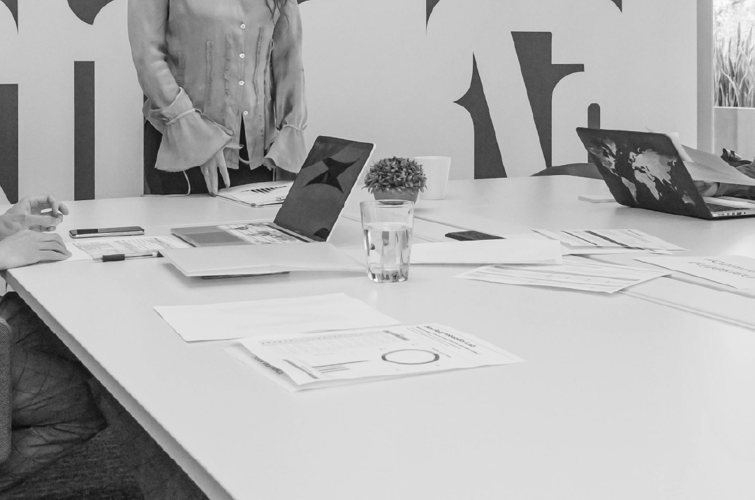 Documents and laptops rest on a large conference room table with two people off to the side.