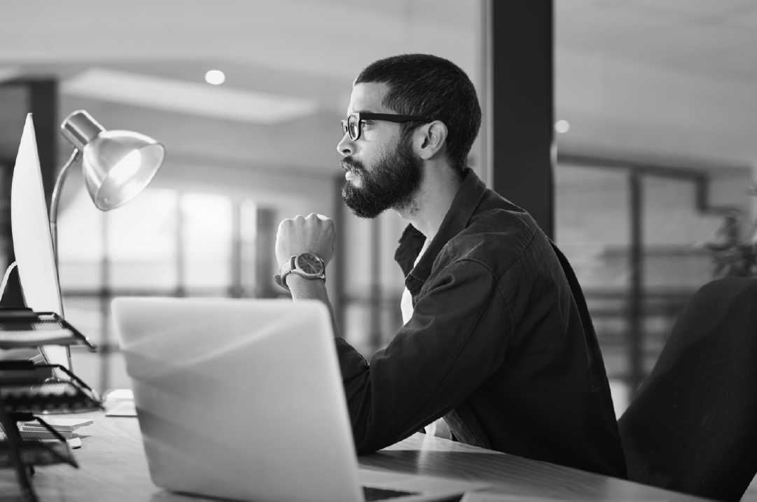 A lone man in an office sits at a desk staring at a monitor next to a desk lamp and a laptop.