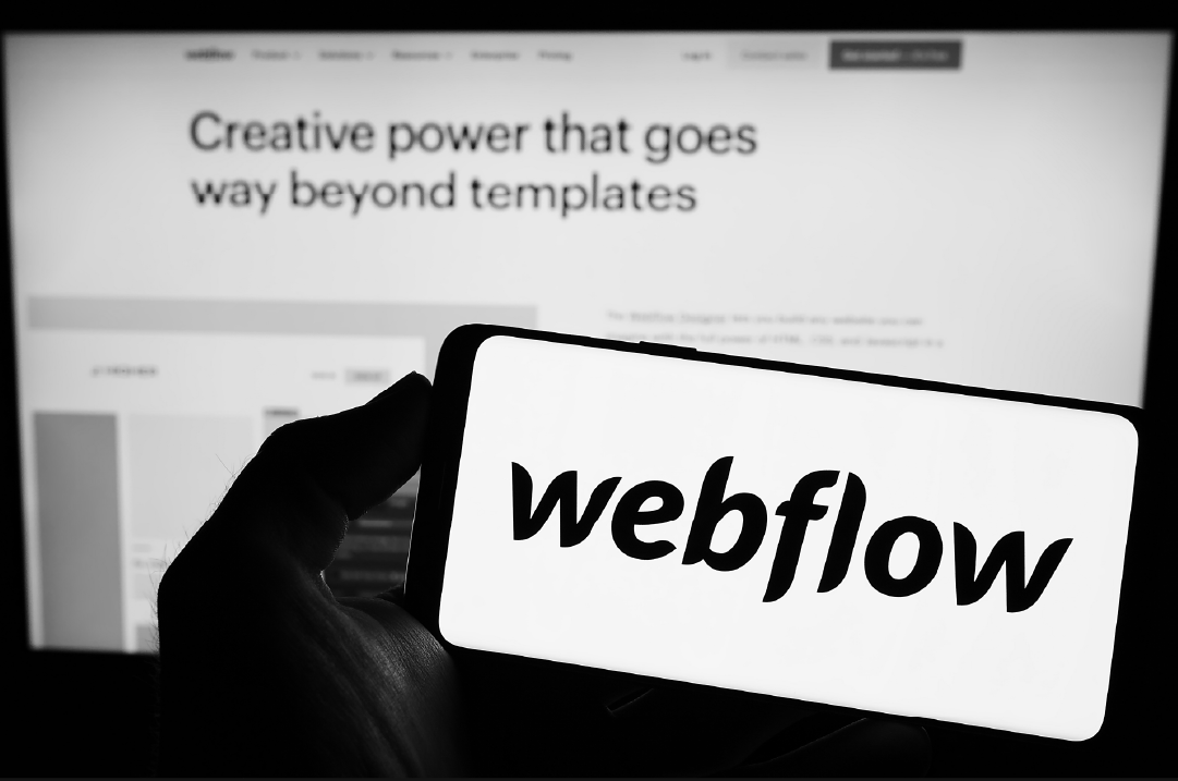 In the foreground, a hand holds a mobile phone that displays the Webflow logo. In the background, a monitor displays a website with the words, "Creative power that goes way beyond templates."