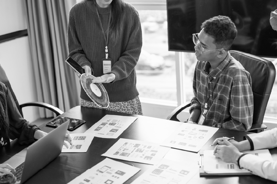 A woman standing in a room holds color swatches in her hand. She is pointing at them while her 3 seated colleagues consider the options.