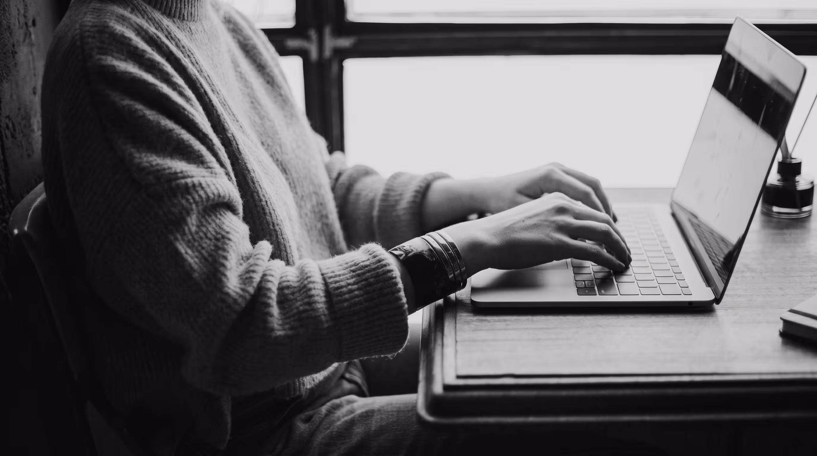 Black and white photo of someone working on a laptop near a window