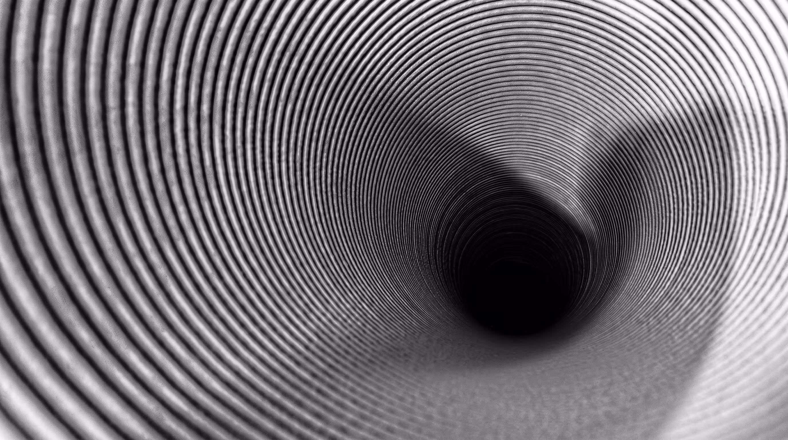 Abstract black-and-white macro photo of a spiral tunnel with concentric ridged rings converging toward a dark center point, creating a symmetrical, hypnotic funnel effect.