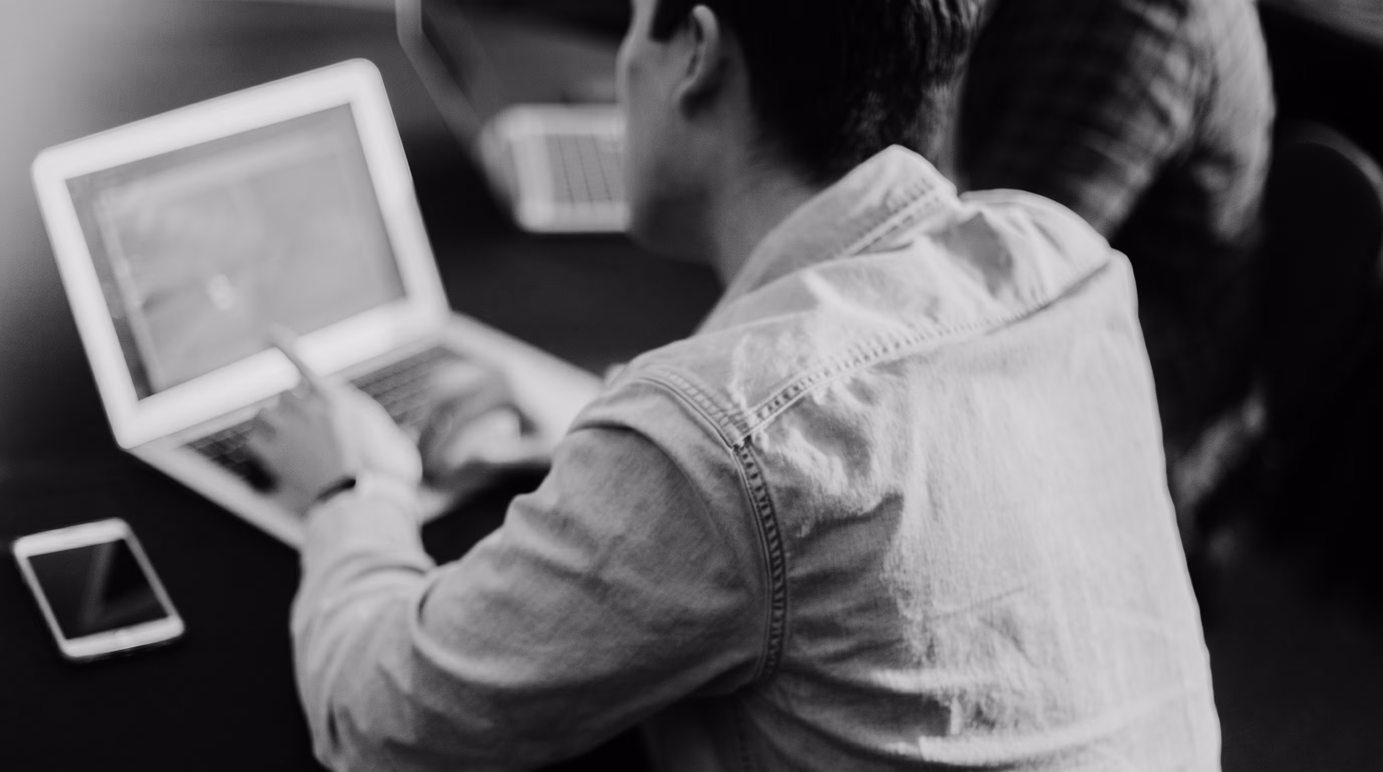 Black-and-white photo taken from over the shoulder of a person typing on a laptop, with a smartphone on the desk nearby and another laptop visible in the background.