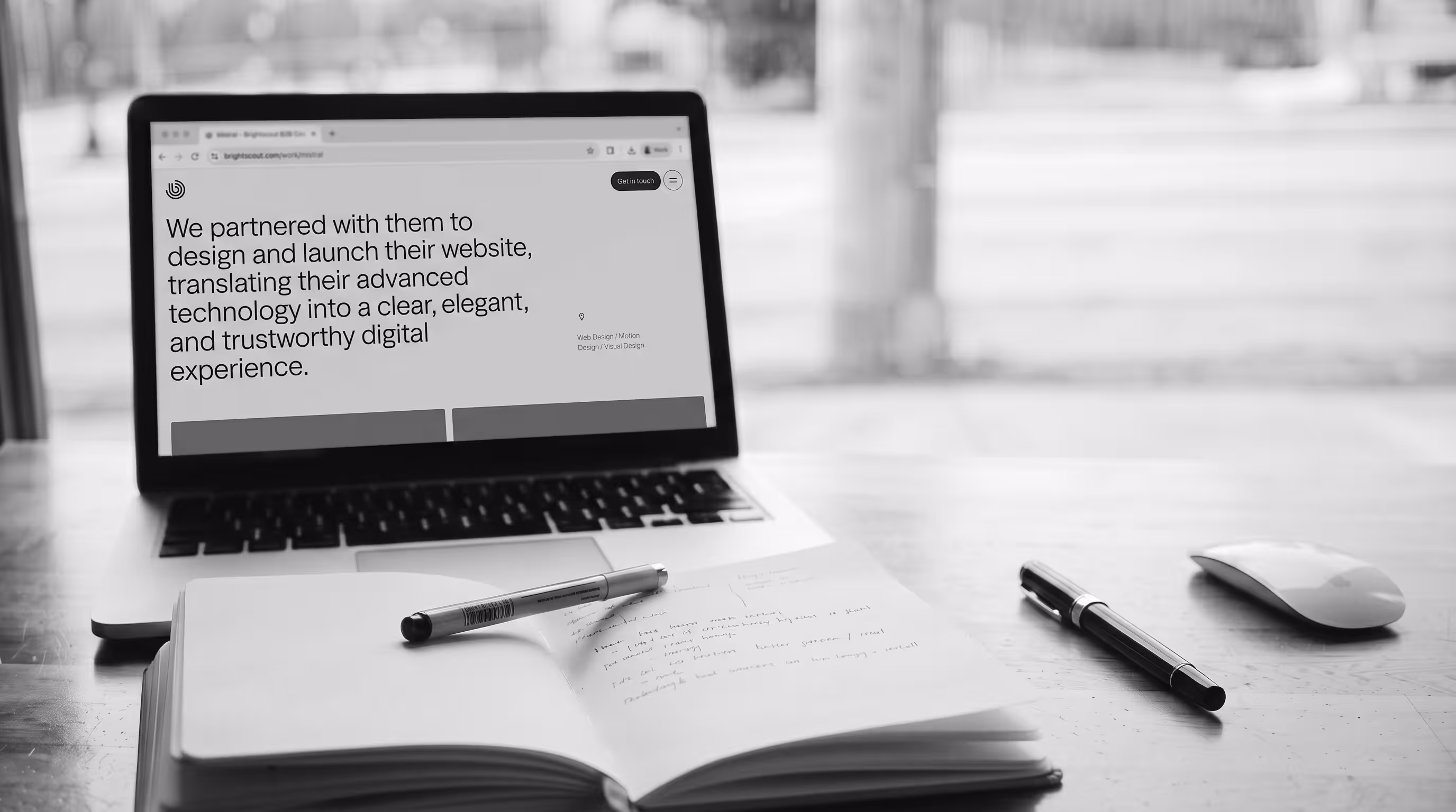 Black and white desk scene with a laptop open to a case study and a notebook full of handwritten notes