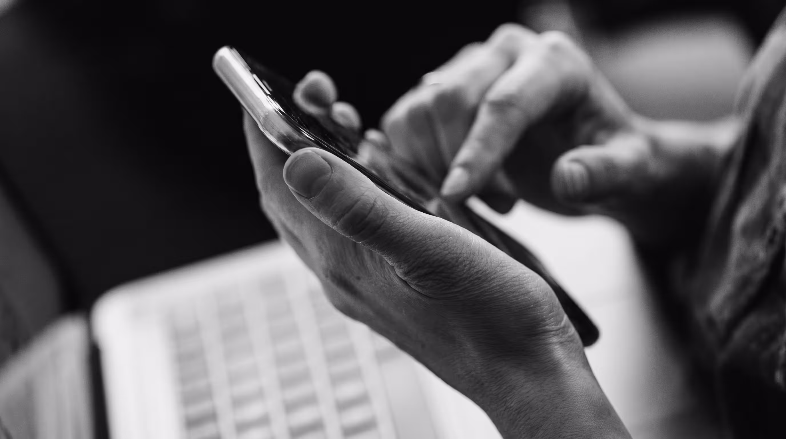Black and white close-up of a person using a phone at a desk with a laptop behind it