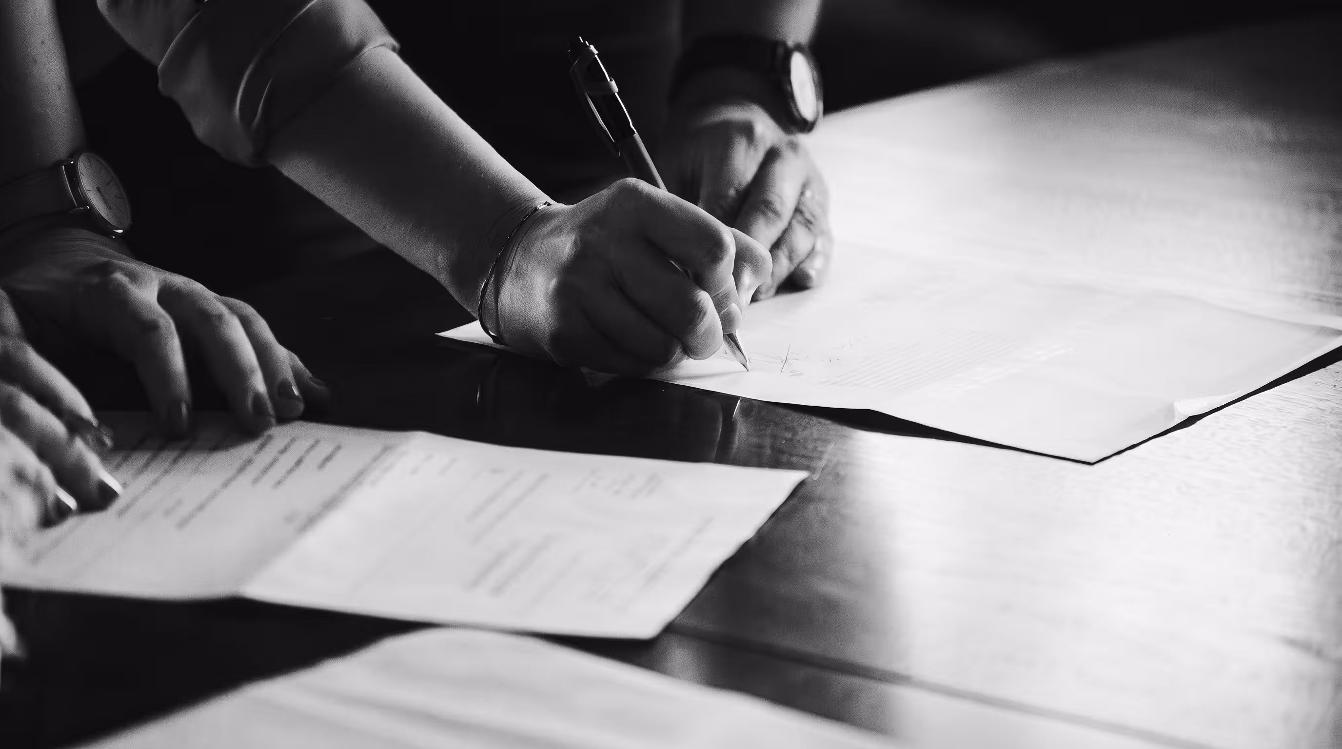 Black and white photo of people gathered over a contract, one hand signing