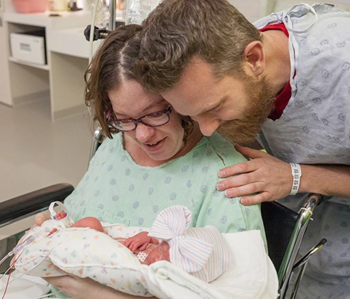 A woman in a hospital gown and wheelchair hugs her newborn baby while her husband peers over her shoulder