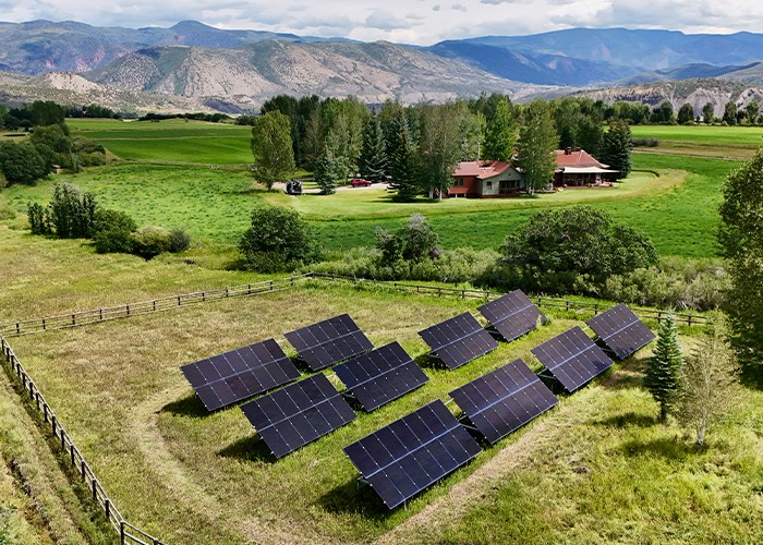 Ground-mounted solar panels in a grass field in front of a ranch house with mountains behind