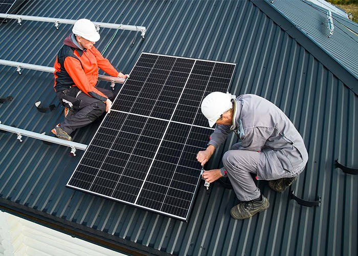 Two workers in hard hats installing a solar panel on a dark metal roof